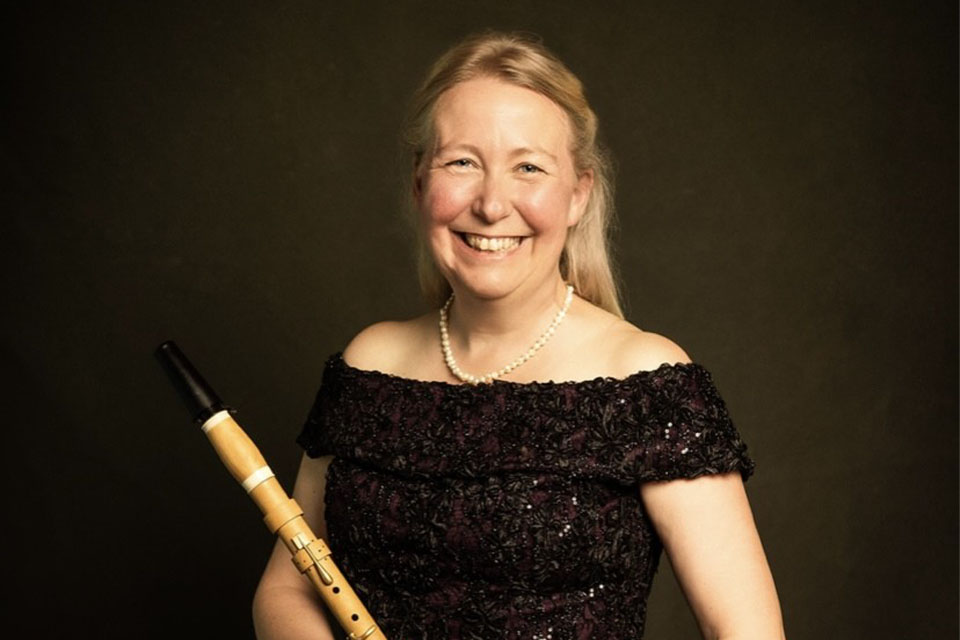 A white woman with blonde hair smiling, wearing a dark sequined top and a necklace. She is holding a historical clarinet.